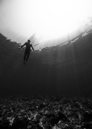 Black and white of from below of scuba diver swimming in clear water in swimsuit under rays of light